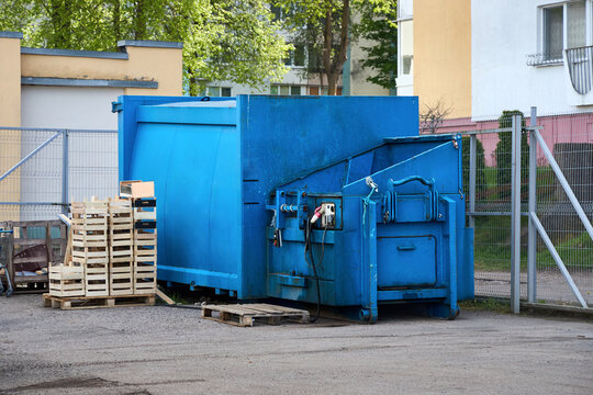 Large blue industrial waste compactor sits on asphalt near wooden pallets and crates. Metal fence and residential building with trees appear in background. Heavy machinery used for urban refuse.