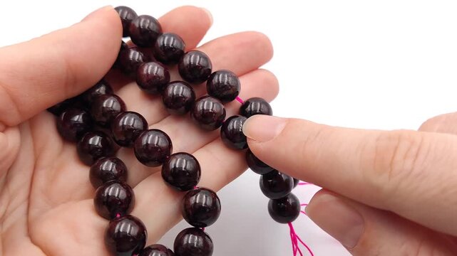 Natural deep wine red garnet crystal beads are closely held and inspected in two parallel rows on a thin pink thread, showcasing their glassy reflective surfaces and rich burgundy undertones