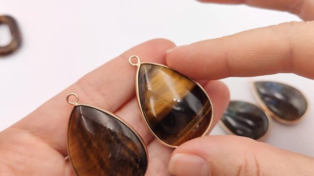 Two large, teardrop-shaped tigers eye gemstone pendants are held and turned in an open hand, showcasing their rich golden-brown bands, chatoyant light reflection, and smooth, polished surfaces