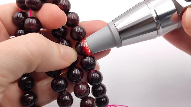 A person uses a powerful flashlight to test the authenticity of deep red garnet gemstone beads on a pink string showing rich internal color variations and natural clarity under bright light