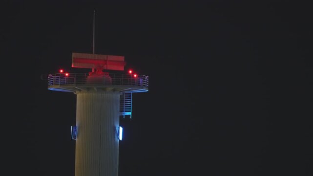 Airport surveillance radar tower photographed at night, lit by red obstruction warning lights and blue ambient LEDs, with the rotating radar antenna standing out against the dark sky