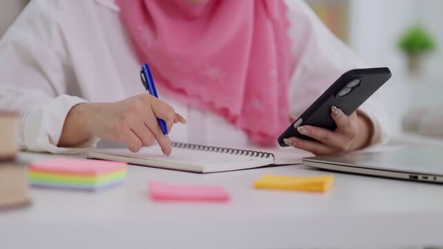 Muslim businesswoman hands using smartphone writing notes planning business tasks with pen notebook working from home office desk remote work multitasking