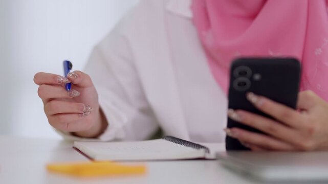 Muslim business women multitask mobile smartphone check message text notebook write notes pen hand casual office hijab pink modern workspace desk organize