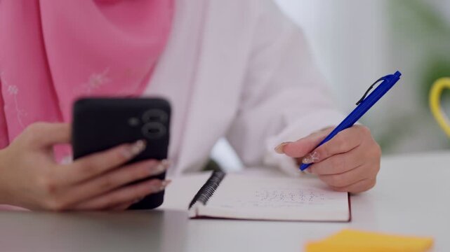 Muslim businesswoman writing notes in notebook while checking smartphone multitask planning schedule workflow task management at desk workspace home office