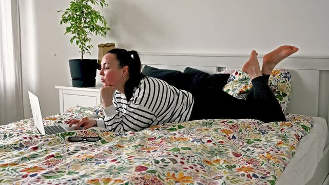 Woman casually browsing laptop in relaxed position on bed in cozy bedroom with plants and white interior, comfortable evening leisure and digital entertainment at home