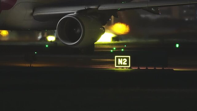 Close-up of a commercial jet engine and wing at night, dramatically lit by glowing yellow runway lights and green taxiway centerline lights with an illuminated taxiway sign in the foreground