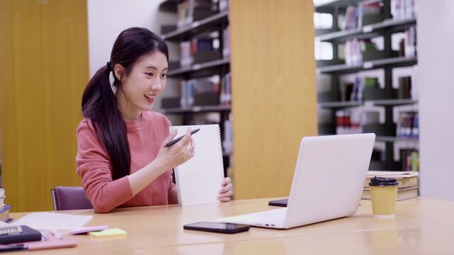 Asian student young women studying notebook writing notes research reading online laptop computer library education modern bookshelf learning online course