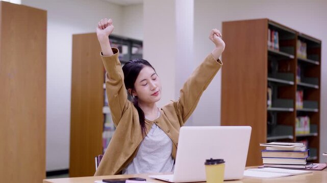successful Asian student stretching arms celebrating achievement victory with laptop computer books stack studying learning research at library desk education