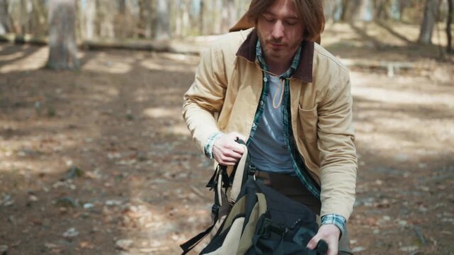 Male tourist opening water bottle during hiking break. Caucasian traveler sitting near backpack after long forest route. Bearded man resting outdoors and preparing for continued woodland expedition.