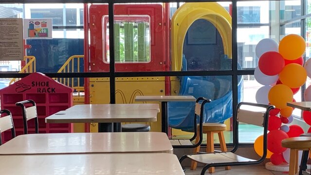 Indoor dining area with tables and chairs facing glass wall, revealing colorful children&rsquo;s playground equipment and decorative balloons