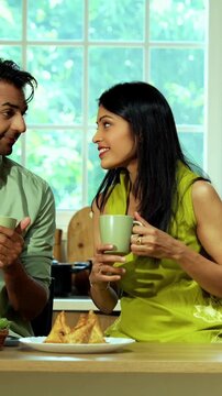 Indian asian young husband and wife having coffee in modern kitchen platform with light hearted chit chat enjoying their morning beverage and talking together in a relaxed home environment