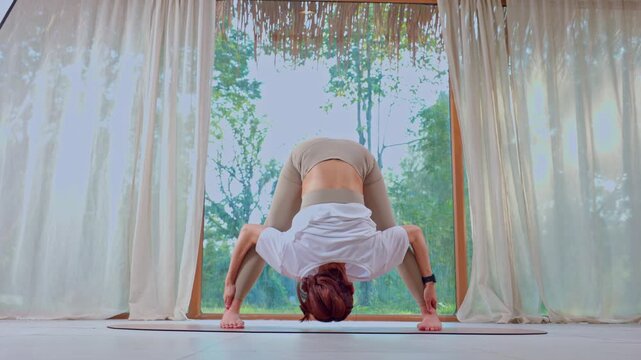 Yoga woman stretch forward bend indoor window home workout, wide legged forward fold on mat by large window, calm wellness fitness flexibility