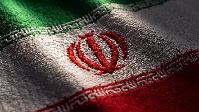 Iran's flag waves softly in close-up. Green, white, red stripes stretch across the frame. Red emblem sits at the center. Soft lighting highlights fabric texture. Gentle motion creates dynamic visuals