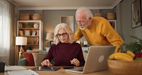 An older couple is at home, focusing on a tablet and a laptop. They discuss life insurance and finances while surrounded by bookshelves and a bowl of fruit. © Stockphotodirectors