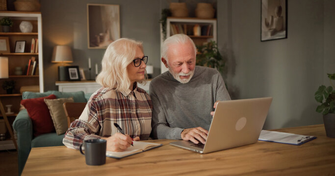 Two older adults sit together at a table in a cozy living room. They focus on a laptop while taking notes and discussing life insurance choices and financial plans.