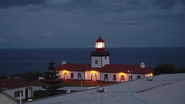 Farol de Albernaz Lighthouse at Twilight with Glowing Lantern and Warm Window Illumination, Historic Maritime Architecture Against Dark Moody Clouds and Atlantic Ocean Horizon, Flores Island, Azores