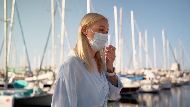 Woman wear surgical mask coughing and show symptoms of respiratory virus like coronavirus or hantavirus while walk in marina with yachts in background