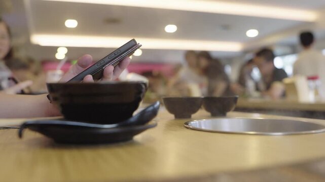 Close-up of hands using a smartphone at a restaurant table with black bowls. Casual dining scene showing digital technology in social settings with a blurred background of people eating.