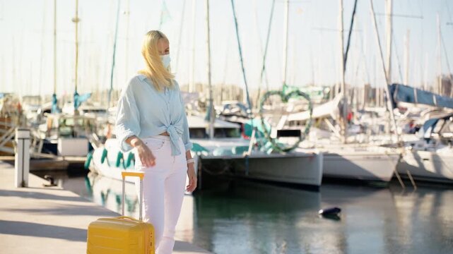 Blonde woman with yellow suitcase wearing protective face mask at marina near yachts, feeling unwell while waiting. Travel safety, health precautions, and cautious journey concept.