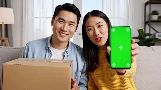Happy Asian couple unboxing package and showing phone with green screen.