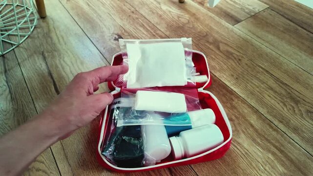 Red first aid kit closure demonstration with hands packing pills, blister packs, bottles and gauze on wooden floor, traveler preparing compact pouch for trip, zipper closing, travel
