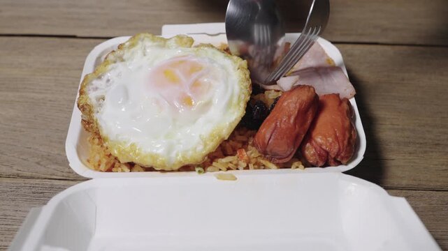 American Fried Rice in a paper takeout container on a wooden table. Includes a sunny-side-up fried egg, small sausages, and sliced ham. Popular Thai street food dish for delivery and quick meals.