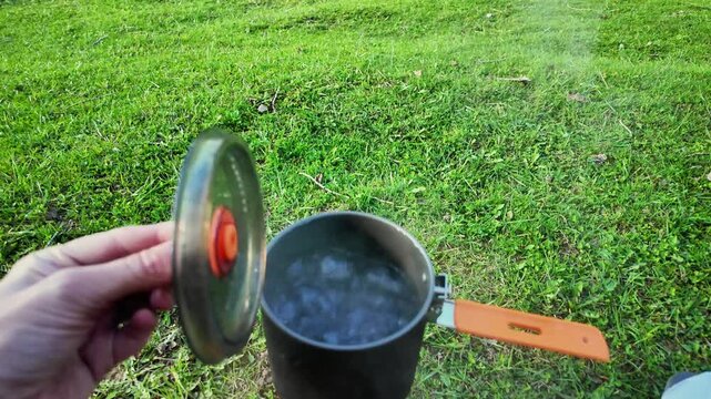 Hand adjusts portable stove valve on grassy field, white traveler primes fuel and checks flame under black pot, picnic bread and cup nearby, efficient outdoor cooking setup