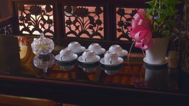 Six white ceramic tea cups are placed upside down on their respective plates near the vase with flowers on a well-polished wooden table prepared for Gongfu Cha (tea art) ceremony 