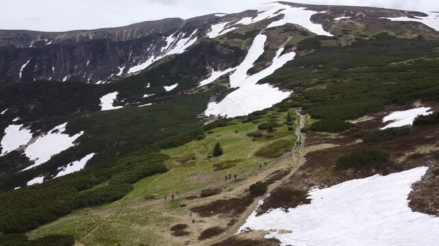 Drone footage of hikers walking on a mountain trail near melting snow patches. Camera captures the rugged spring alpine landscape with green shrubs and steep slopes.