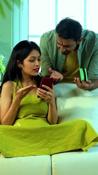 Indian asian young couple duped with online fraud while using electronic credit or debit card for online purchase using smartphone sitting on couch in modern Indian home looking worried