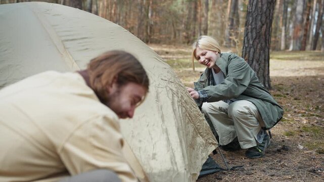 Caucasian couple assembling camping tent in forest area. Smiling woman helping male traveler prepare campsite outdoors. Hikers organizing temporary shelter during woodland adventure vacation.