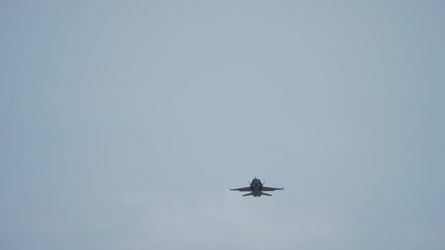 A twin-engine military fighter jet is seen head-on from below in a pale hazy sky, its wide wingspan creating a powerful symmetric silhouette