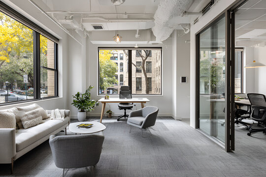 A modern office space, showcasing a bright room with seating areas, a desk, a window overlooking greenery, and an adjacent glass-walled area