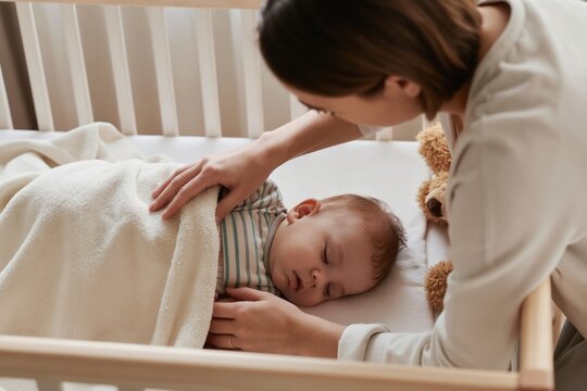 Mother gently tucks sleeping baby into crib. Soft blanket covers infant. Teddy bear nearby. Calm nursery scene, peaceful bedtime moment. New parent love, gentle care.