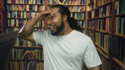 Man in white t shirt shielding eyes with hand while smiling and gesturing toward bookshelves in a building library aisle  surprise discovery learning. © Krakenimages.com