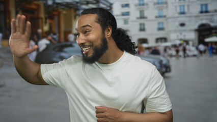 Fototapeta premium Man smiling and waving his bare hand on a busy street by a parked car and building, wearing a white t shirt and ponytail; friendly greeting.