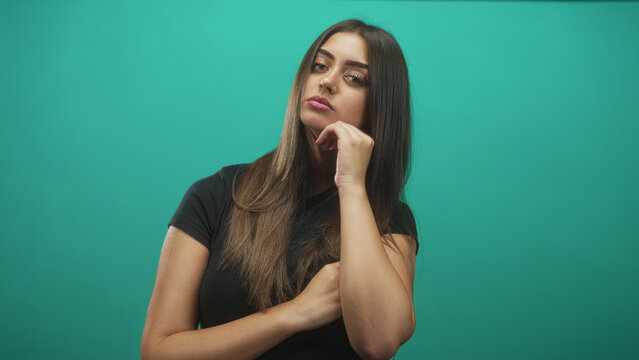Woman touching chin with hand and arm folded holding elbow in studio wearing black shirt against teal backdrop; pensive.