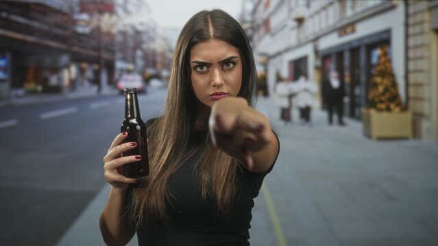 Woman holding beer bottle points finger to camera on a city street with storefronts and christmas tree, visible fingernail and stern gaze; defiance anger.