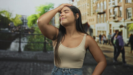 Fototapeta premium Woman touching forehead with raised hand and closed eyes on a street beside a canal and buildings; relief travel memory.