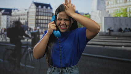 Fototapeta premium Young hispanic woman holding a blue corded telephone to her ear and pressing hand to forehead on a crowded street near steps and a statue; frustration.