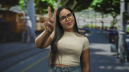 Fototapeta premium Young hispanic woman wearing glasses shows peace sign with right hand and bare midriff visible on a tree lined city street; youthful confidence.