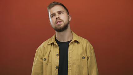 Obraz premium Man tilting head with visible face and beard, wearing yellow jacket against orange studio backdrop; pensive.
