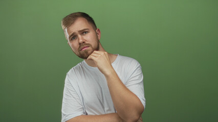 Fototapeta premium Man wearing white tshirt with hand on chin touching beard in green studio; thinking doubt curiosity.