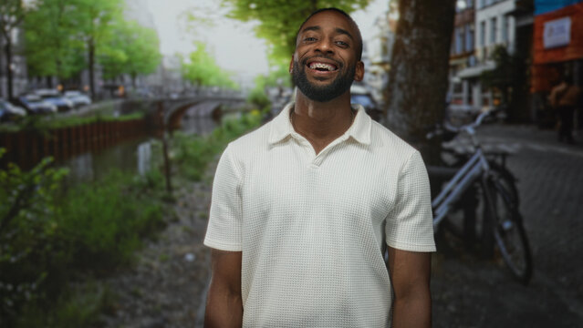 Young man smiling showing teeth and braces on a street by a canal with bicycle and trees in daylight; quiet contentment.