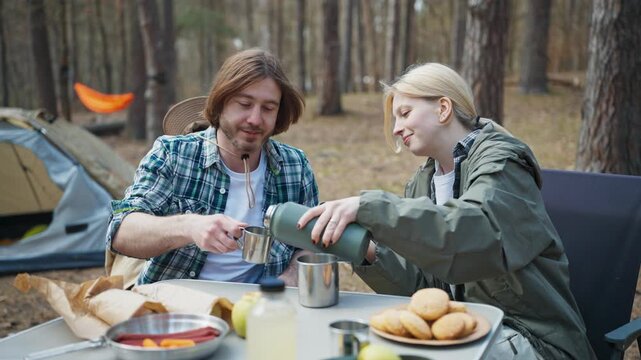 Caucasian couple pouring hot drink into metal cups at forest campsite table. Travelers enjoying outdoor breakfast near tent during camping trip and hiking adventure in woodland nature.