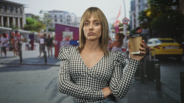Woman holds takeaway coffee cup with crossed arm and raised wrist, neutral unimpressed expression on a busy city street; indifference.