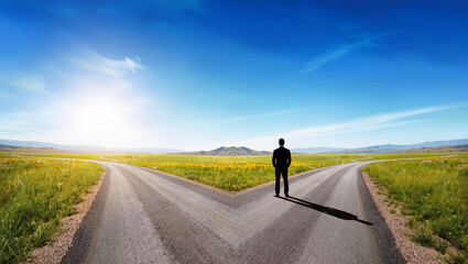 Fototapeta premium Businessman standing at crossroads facing two diverging roads in vast countryside landscape, conceptual decision making and choice metaphor, editorial business strategy photography with copy space