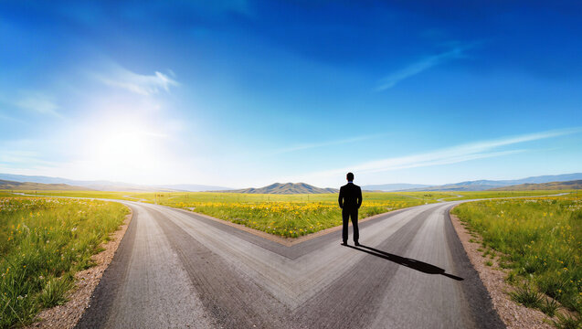 Businessman standing at crossroads facing two diverging roads in vast countryside landscape, conceptual decision making and choice metaphor, editorial business strategy photography with copy space