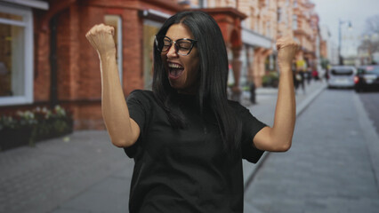 Fototapeta premium Woman in glasses wearing black tshirt raises clenched fists and bites lower lip on street with brick storefronts and parked cars; victory joy.
