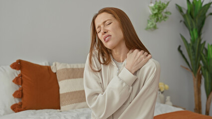 Naklejka premium Woman holding shoulder wincing on bed in building bedroom, eyes closed and hand on shoulder; discomfort recovery.